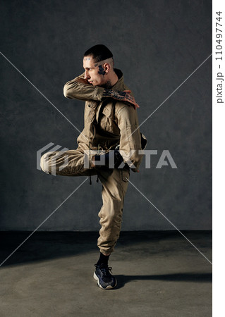 Handsome male with tattooed body and face, earrings, beard. Dressed in khaki jumpsuit, black sneakers. Dancing on gray background. Dancehall, hip-hop 110497744