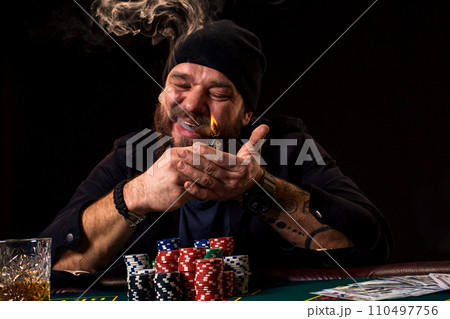 Bearded man with cigar and glass sitting at poker table in a casino. Gambling, playing cards and roulette. 110497756