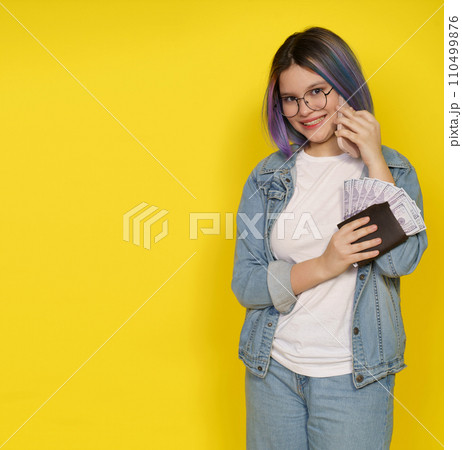 Financial Independence Associated With Managing Money In Modern And Confident Manner. Happy Smiling Teenager Girl With Wallet Full Of Money And Mobile Phone Isolated On Yellow With Copy Space. High 110499876