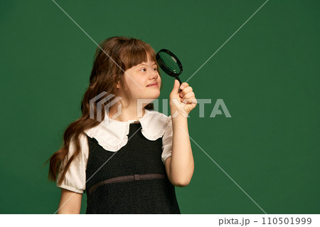 Teen girl with down syndrome looking in magnifying glass against green studio background. Education for people with special needs Teen girl with down syndrome looking in magnifying glass against green studio background. Education for people with special needs 110501999