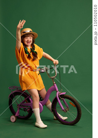 Happy teen girl with down syndrome wearing yellow suit, sitting on bicycle and having fun against green studio background Happy teen girl with down syndrome wearing yellow suit, sitting on bicycle and having fun against green studio background 110502010