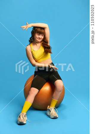 Sport therapy. Teen girl with down syndrome sitting on fitness ball and training against blue studio background 110502025
