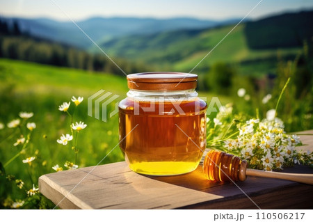 Jar of honey and chamomile on a wooden table with green fields in the background 110506217