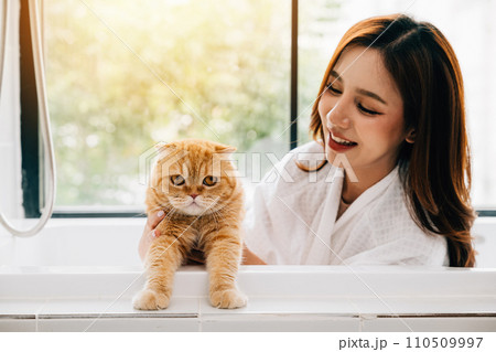 Bathing bliss, an adult woman smiles while holding her Scottish Fold cat in the bathroom, creating a scene of relaxation and pet adoration. Bathing bliss, an adult woman smiles while holding her Scottish Fold cat in the bathroom, creating a scene of relaxation and pet adoration. 110509997