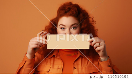 Portrait of beautiful young woman with red curly hair in orange coat holding blank card over her face. 110510192