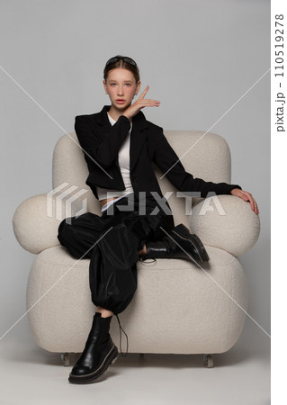 A beautiful slender girl in a black suit sits in a cozy white chair on a gray background. 110519278