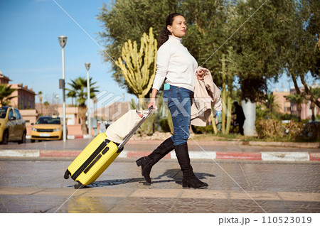Woman traveler with yellow suitcase, crossing the street on pedestrian crosswalk, confidently looking into the distance. 110523019