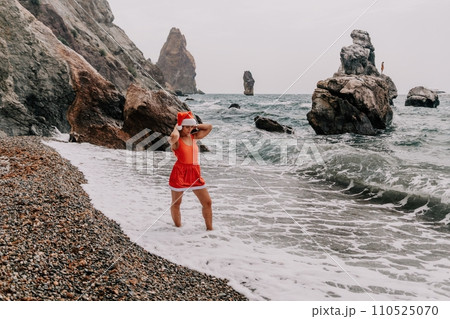 Woman summer travel sea. Happy tourist in red bikini and Santas hat enjoy taking picture outdoors for memories. Woman traveler posing on the beach surrounded by volcanic mountains, sharing travel joy 110525070