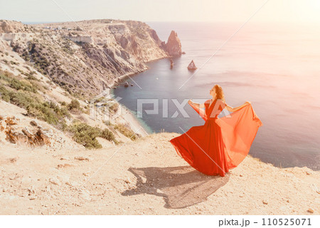 Woman in red dress on sea. Side view a Young beautiful sensual woman in a red long dress posing on a rock high above the sea on sunset. Girl on the nature on blue sky background. Fashion photo. 110525071