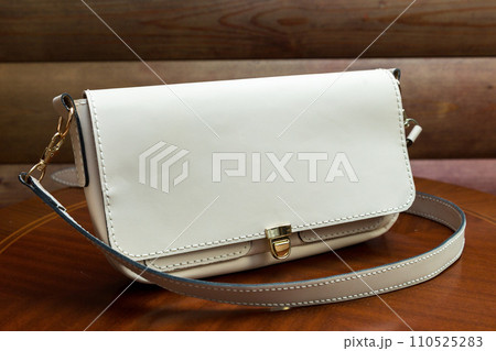 White leather women's handbag with a strap on wood background. An elegant accessory for stylish women.. .Handbag made of genuine leather, comfortable and practical. 110525283