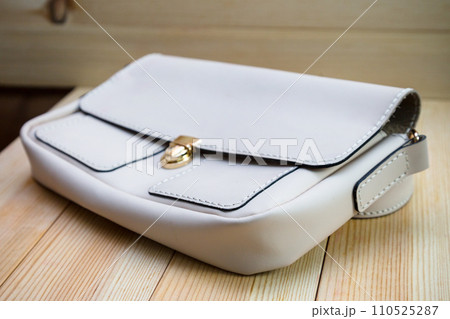 White leather women's handbag with a strap on a light wood background. An elegant accessory for stylish women.. .Handbag made of genuine leather, comfortable and practical. 110525287