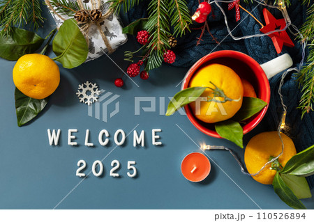 Welcome 2025 text. Winter composition with fir branches, present, cones and tangerines on a blue background. Cozy winter mood. View from above. Welcome 2025 text. Winter composition with fir branches, present, cones and tangerines on a blue background. Cozy winter mood. View from above. 110526894
