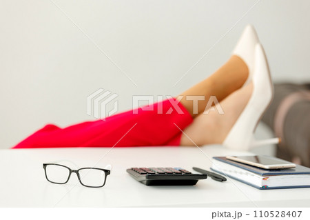 Cropped shot of businesswoman's legs in red pants and white high heels shoes on office desk relaxing during break at work. Selective focus on eyeglasses, calculator, notebook and smartphone on table Cropped shot of businesswoman's legs in red pants and white high heels shoes on office desk relaxing during break at work. Selective focus on eyeglasses, calculator, notebook and smartphone on table 110528407