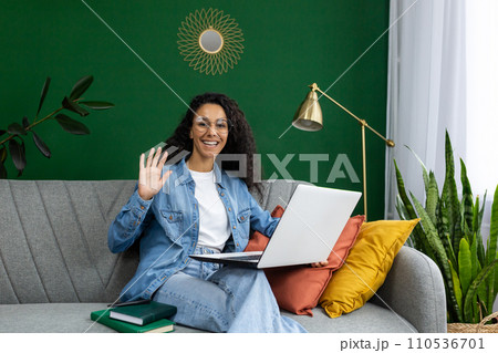 Cheerful woman with glasses waving while holding a laptop, sitting on a couch with colorful cushions and indoor plants. Cheerful woman with glasses waving while holding a laptop, sitting on a couch with colorful cushions and indoor plants. 110536701