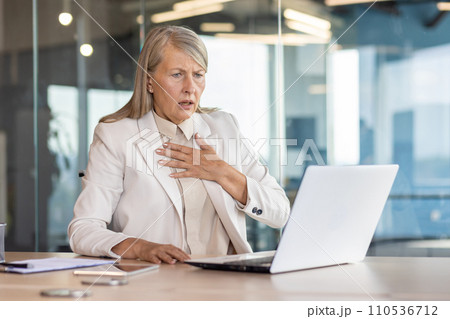 Senior woman in business suit sitting at desk in office in front of laptop and holding hand to chest, having panic attack, heart attack, shocked by business affairs. Senior woman in business suit sitting at desk in office in front of laptop and holding hand to chest, having panic attack, heart attack, shocked by business affairs. 110536712