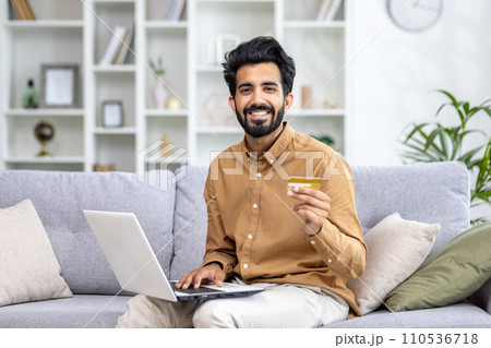 Smiling man with beard shopping online using laptop and credit card on cozy sofa, home interior background, e-commerce concept 110536718