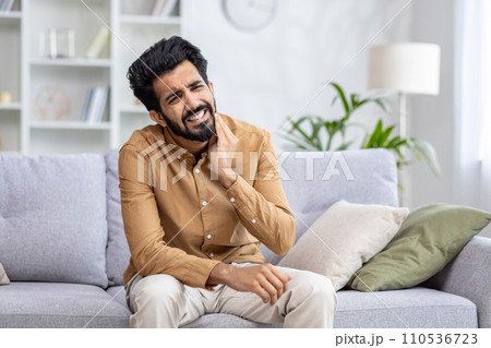 Man in pain, holding cheek, suffering toothache, sitting on couch at home, expressing discomfort and agony, dental problem concept. 110536723