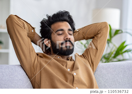 Relaxed bearded man with closed eyes resting on a couch, hands behind head in a cozy room, concept of peace and leisure time at home. Relaxed bearded man with closed eyes resting on a couch, hands behind head in a cozy room, concept of peace and leisure time at home. 110536732