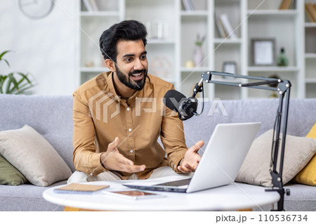 Enthusiastic man podcasting with laptop and microphone at home, expressing positivity and engagement in a cozy setting. 110536734
