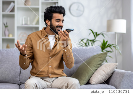 Joyful man speaking into smartphone like a microphone on a cozy sofa, expressing happiness and comfort in a modern living room. Joyful man speaking into smartphone like a microphone on a cozy sofa, expressing happiness and comfort in a modern living room. 110536751
