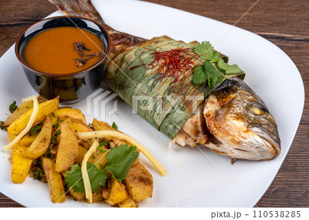 Baked dorado fish in bamboo leaves sprinkled with cilantro and chili threads, baked potatoes and Szechuan sauce. Baked dorado fish in bamboo leaves sprinkled with cilantro and chili threads, baked potatoes and Szechuan sauce. 110538285