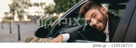 Smiling male driver in suit is riding behind steering wheel of car and looks camera 110538715