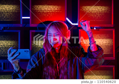 Winning. Confused Asian woman wearing gaming headphones feeling excited at gaming room, Happy Gamer playing video game online with smart mobile phone with neon lights raises hands to wins celebrating Winning. Confused Asian woman wearing gaming headphones feeling excited at gaming room, Happy Gamer playing video game online with smart mobile phone with neon lights raises hands to wins celebrating 110540620