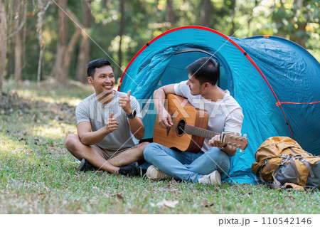 LGBTQIA Gay couple camping together in woods for holidays and relax on guitar together LGBTQIA Gay couple camping together in woods for holidays and relax on guitar together 110542146