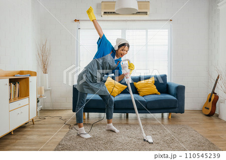 Happy young Asian housewife sings her favorite song with a vacuum cleaner as a microphone while cleaning. Enjoying domestic work dancing in living room a cheerful maid having fun. Happy young Asian housewife sings her favorite song with a vacuum cleaner as a microphone while cleaning. Enjoying domestic work dancing in living room a cheerful maid having fun. 110545239
