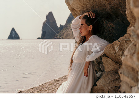 Woman summer travel sea. Happy lady with long hair in white dress enjoy taking photo outdoors for memories. Woman traveler posing on beach at sea surrounded by volcanic mountains, sharing emotions 110548657