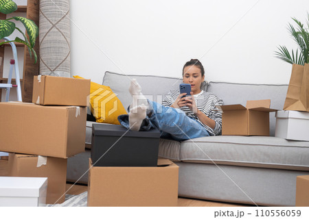 Attractive brunette lady in casual clothes sits on cozy sofa with decorative pillows next to supplies for moving into new space communicating online with friend 110556059
