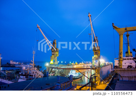 Floating dock in shipyard at dusk, vessels undergoing repair, industrial cranes tower above. Marine maintenance, engineering, shipbuilding, and blue hour add appeal for varied customers. Floating dock in shipyard at dusk, vessels undergoing repair, industrial cranes tower above. Marine maintenance, engineering, shipbuilding, and blue hour add appeal for varied customers. 110556473