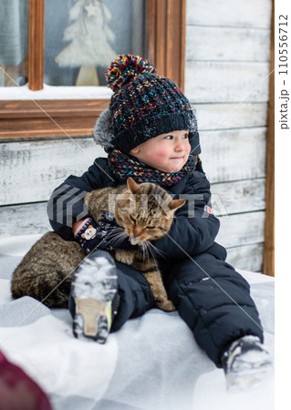 A little boy sits in the snow and holds a cat 110556712