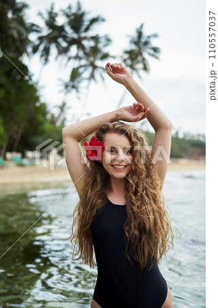 Woman with red hibiscus in hair enjoys tropical sea. Female in black swimsuit at beach escape. Solo traveler explores coast, enjoys summertime swim. Wellness retreat in island paradise. 110557037