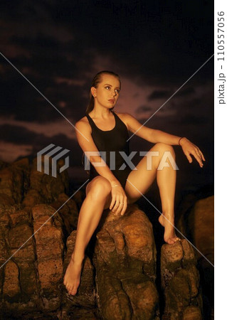 Elegant woman models in black swimsuit on rocky beach at dusk. Athletic female poses, serene seascape background. Swimwear fashion shoot, outdoor lifestyle, summer evening allure. Elegant woman models in black swimsuit on rocky beach at dusk. Athletic female poses, serene seascape background. Swimwear fashion shoot, outdoor lifestyle, summer evening allure. 110557056