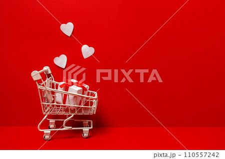 Shopping trolley with gift box, white love hearts on red background. St. Valentine's Day shopping and sale. copy space Shopping trolley with gift box, white love hearts on red background. St. Valentine's Day shopping and sale. copy space 110557242