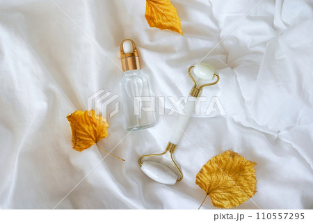 Glass bottle with oil, Gua sha stone for face massage and autumn leaves on white bed background. Cosmetic facial skin care and spa. Natural treatment concept Glass bottle with oil, Gua sha stone for face massage and autumn leaves on white bed background. Cosmetic facial skin care and spa. Natural treatment concept 110557295