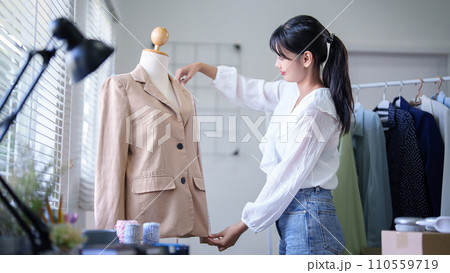 Young asian merchant women using measure tape to checking length size details of fashion clothes in mannequin while taking notes in clipboard and working about online shopping business in home office 110559719