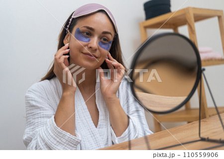 Morning Ritual A contented woman, gazing in the mirror, applies under eye masks to combat puffiness, wrinkles, and dark circles the magic of eye patches.  110559906