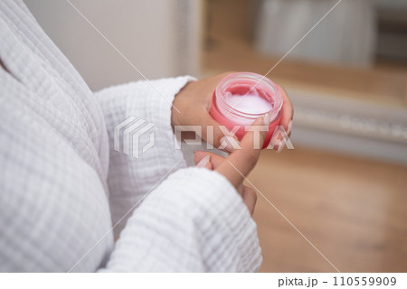 Woman in bathrobe holding open bottle of moisturizing cream near mirror in bathroom lady showing organic cosmetic product for skin pampering in bathroom closeup 110559909