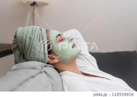 Relaxed young woman with peeling clay mask on face lying on comfortable sofa pretty lady in bathrobe resting during spa procedures with natural cosmetics Relaxed young woman with peeling clay mask on face lying on comfortable sofa pretty lady in bathrobe resting during spa procedures with natural cosmetics 110559916