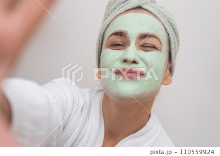 Clay Mask Cheer: A bathrobe-clad woman captures her joyful essence in a selfie with a clay mask, beaming at the camera. Clay Mask Cheer: A bathrobe-clad woman captures her joyful essence in a selfie with a clay mask, beaming at the camera. 110559924