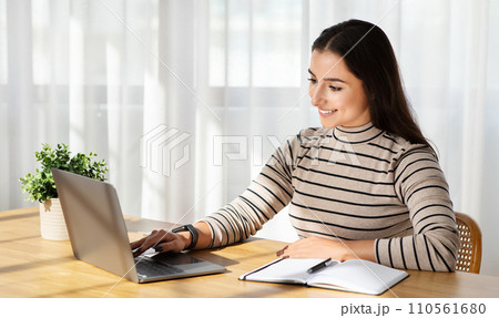 Focused woman with a pleasant smile working on her laptop at a wooden table 110561680