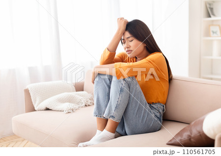 Upset young asian woman sitting on couch, touching her head Upset young asian woman sitting on couch, touching her head 110561705