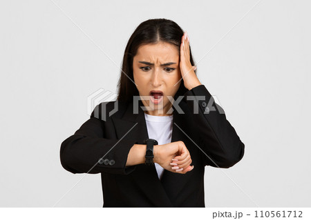 Shocked businesswoman in suit, looking at clock with open mouth and touching head, isolated on gray studio background 110561712