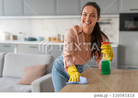 Smiling young woman wiping down table with cleaning spray and cloth 110561818