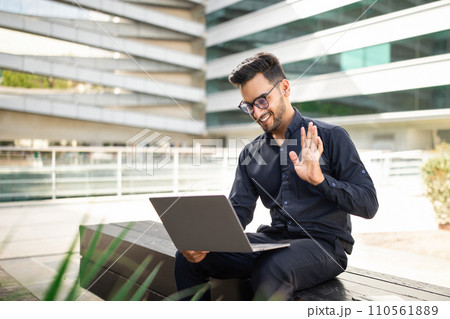 Professional businessman waves hello during video call on laptop outside 110561889