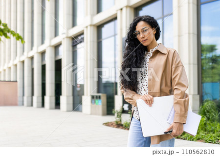 Confident young businesswoman with laptop and notebook standing in front of an office building, exuding professionalism and ambition. 110562810
