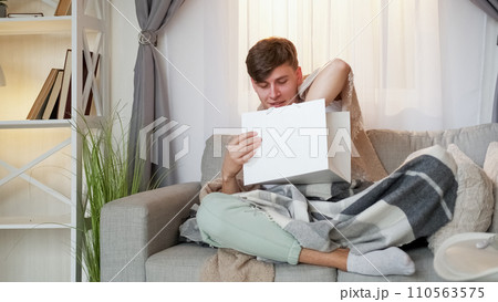 Order receiving. Sickness distance. Home delivery. Smiling ill man with fever wrapped in blanket sitting on sofa with package in living room interior. Order receiving. Sickness distance. Home delivery. Smiling ill man with fever wrapped in blanket sitting on sofa with package in living room interior. 110563575