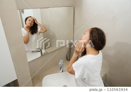 Happy young woman touching her face, examining flaws, skin problems and wrinkles, standing in front of a bathroom mirror 110563934